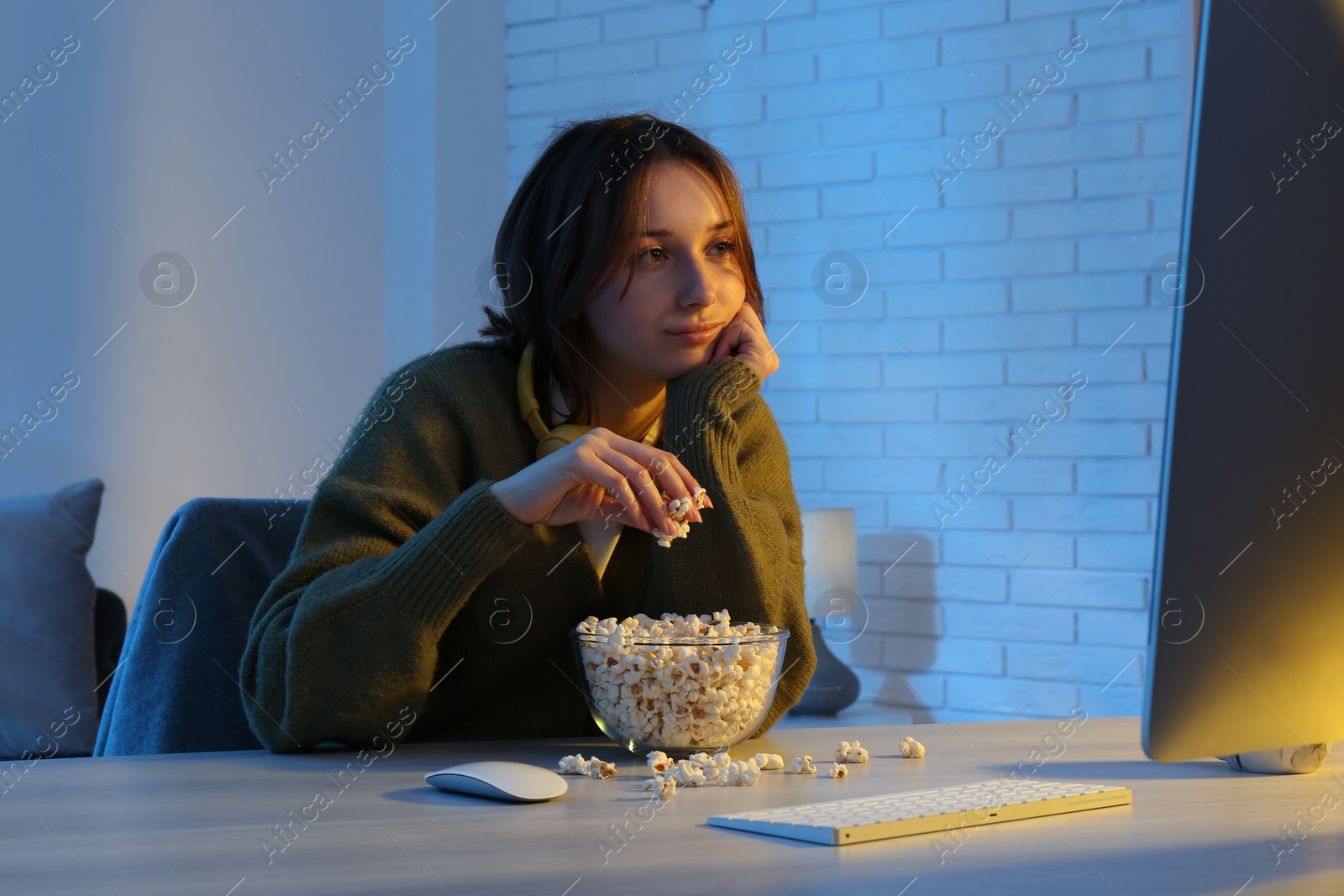 Photo of Beautiful young woman eating popcorn while watching film on computer at table indoors at night