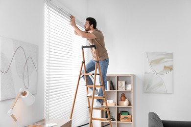 Photo of Man on wooden folding ladder installing blinds at home