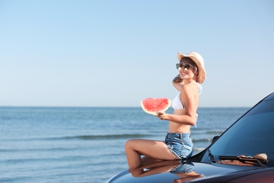 Young woman with watermelon slice near car on beach. Space for text