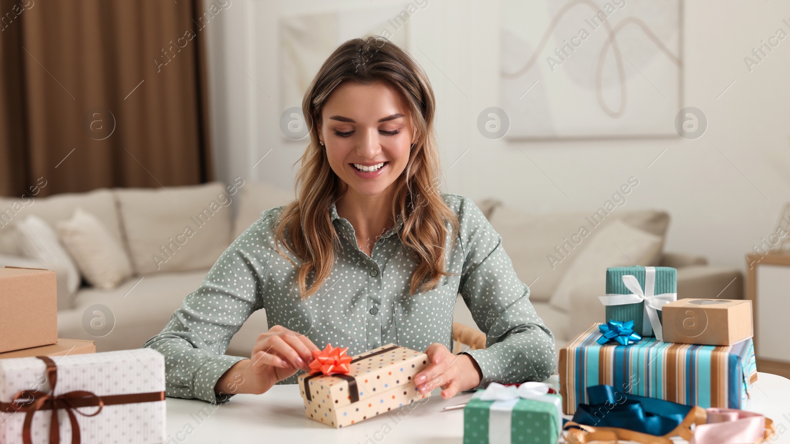 Photo of Beautiful young woman decorating gift box with bow at table in living room