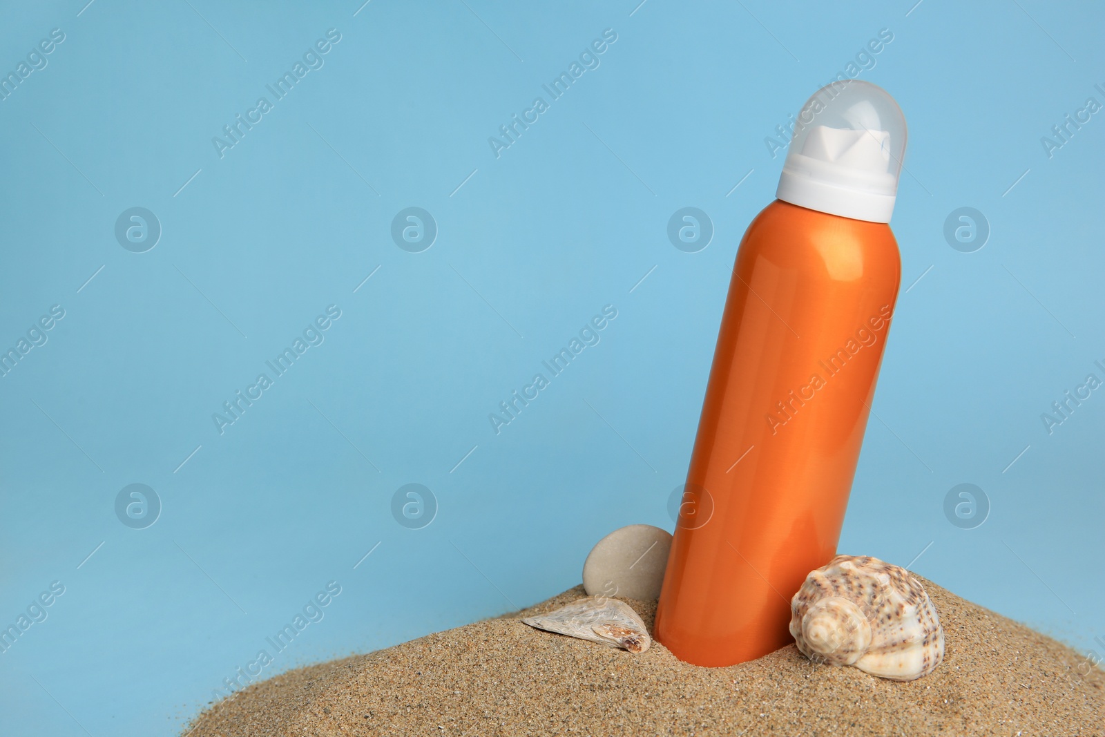 Photo of Sand with bottle of sunscreen, stone and seashell against light blue background, space for text. Sun protection