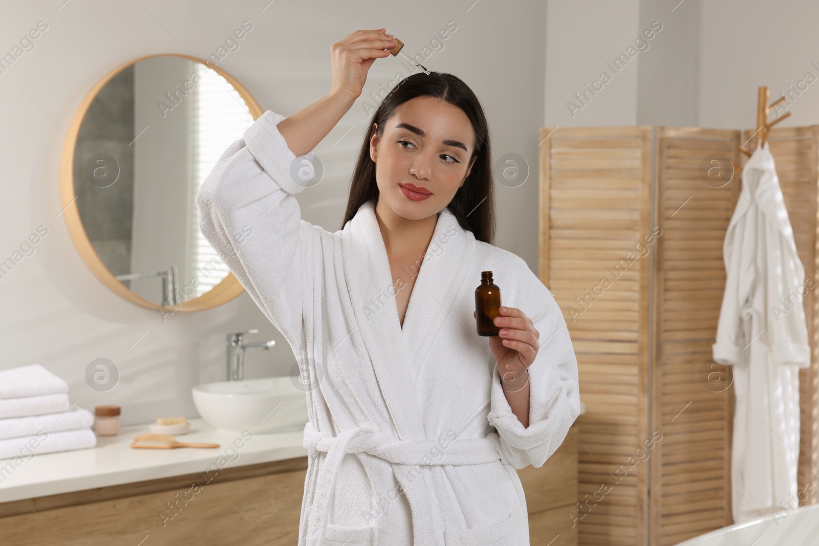 Photo of Happy young woman applying essential oil onto hair roots in bathroom