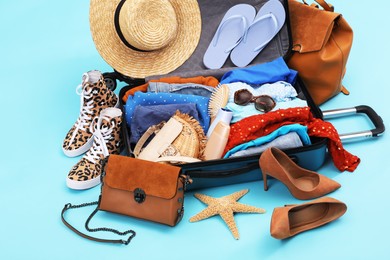 Photo of Open suitcase with clothes, beach accessories and shoes on light blue background. Summer vacation