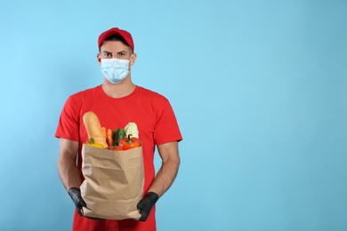 Photo of Courier in medical mask holding paper bag with food on light blue background, space for text. Delivery service during quarantine due to Covid-19 outbreak