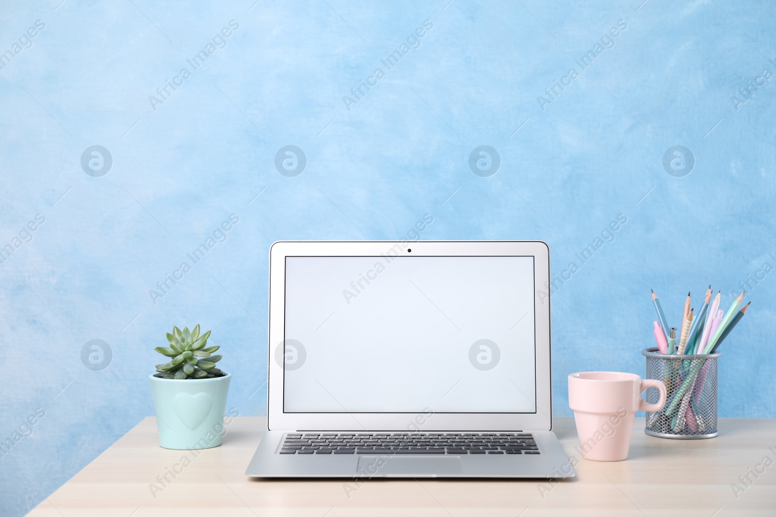 Photo of Modern laptop with blank screen, cup and office supplies on white table near light blue wall. Space for text