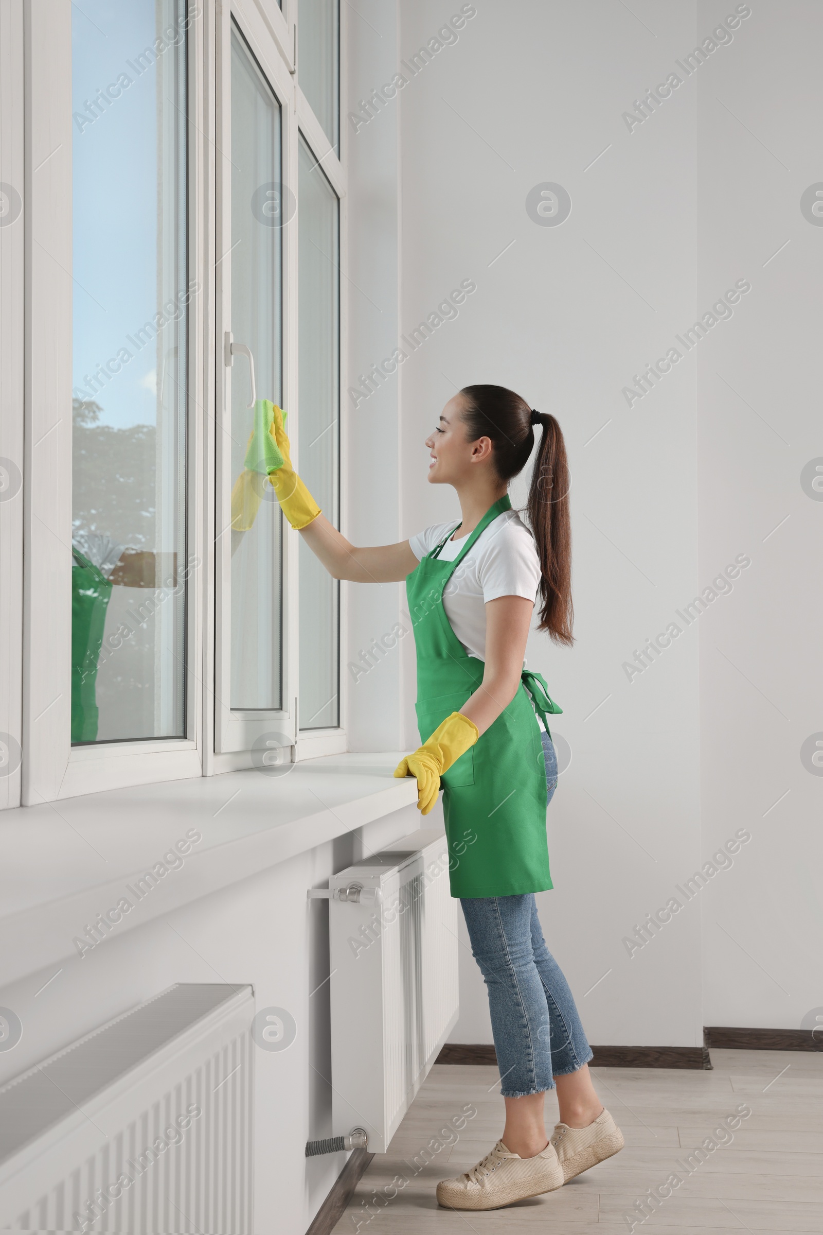 Photo of Happy young woman cleaning window glass with rag indoors