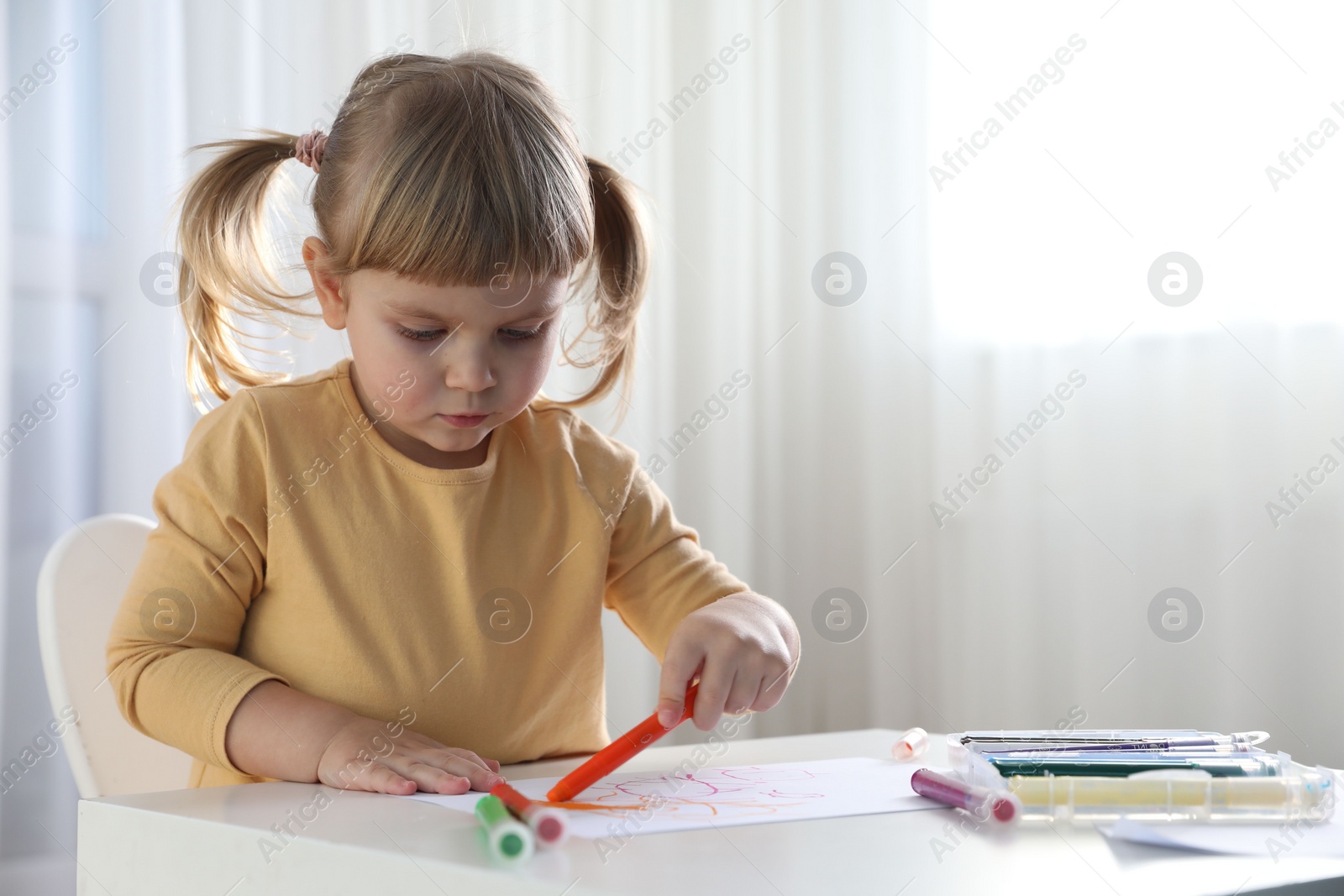 Photo of Cute little girl drawing with marker at white table indoors. Child`s art