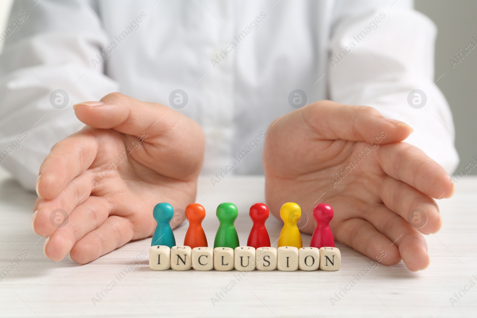 Photo of Woman protecting colorful pawns and wooden cubes with word Inclusion at white table, closeup