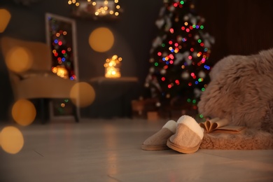 Photo of Soft slippers with fluffy blanket and Christmas tree in dark room