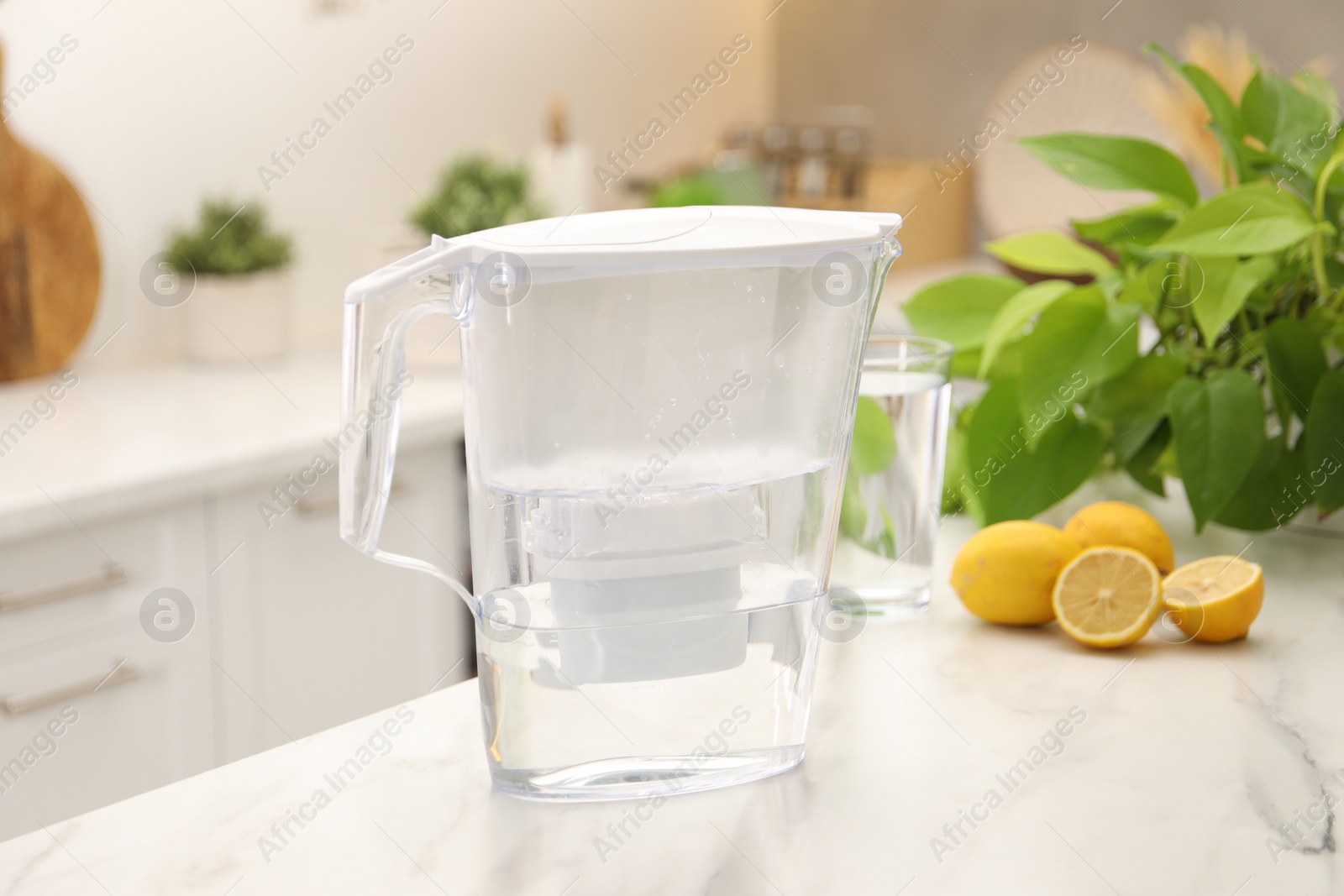 Photo of Water filter jug, glass and lemons on white marble table in kitchen