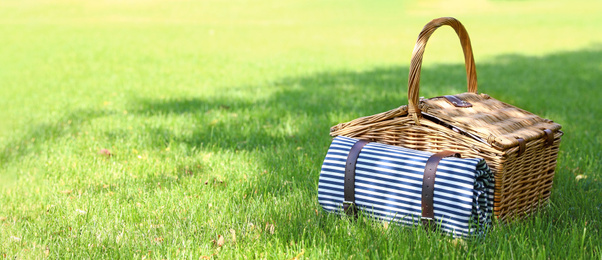 Picnic basket with blanket on green lawn in garden, space for text. Banner design