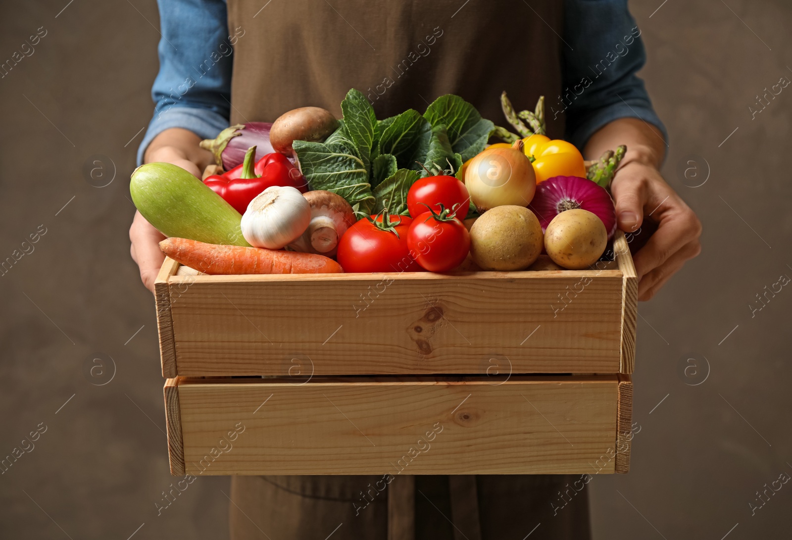 Photo of Man holding wooden crate full of fresh vegetables on brown background, closeup