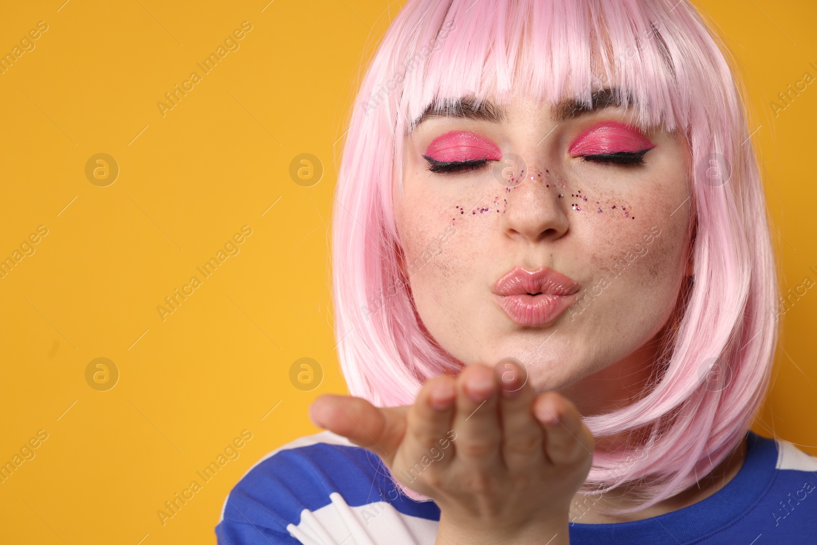 Photo of Beautiful woman with bright makeup and glitter freckles blowing kiss on yellow background, closeup. Space for text