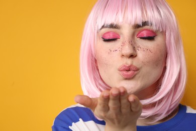 Beautiful woman with bright makeup and glitter freckles blowing kiss on yellow background, closeup. Space for text