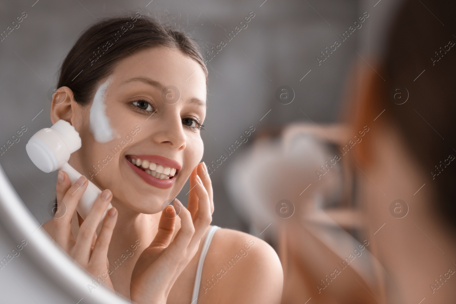 Photo of Young woman washing face with brush and cleansing foam near mirror in bathroom