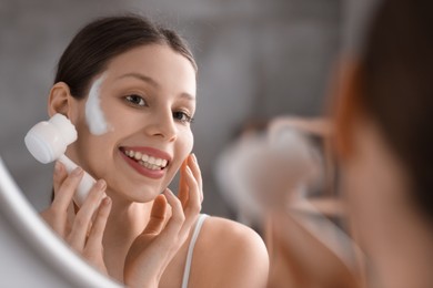 Young woman washing face with brush and cleansing foam near mirror in bathroom