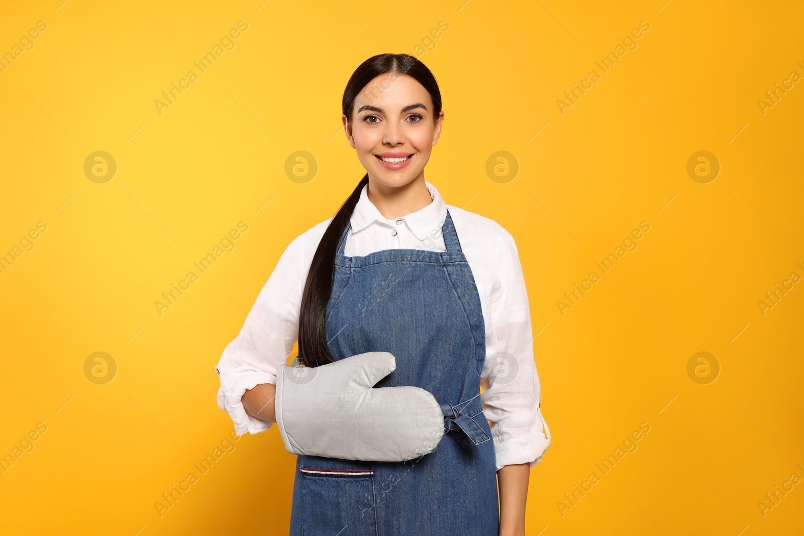 Photo of Young woman in blue jeans apron and oven glove on yellow background