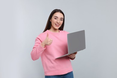 Smiling young woman with laptop showing thumbs up on grey background, space for text