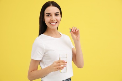 Young woman with glass of water and vitamin capsule on yellow background