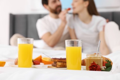 Photo of Tray with tasty breakfast on bed. Husband feeding his wife in bedroom, selective focus