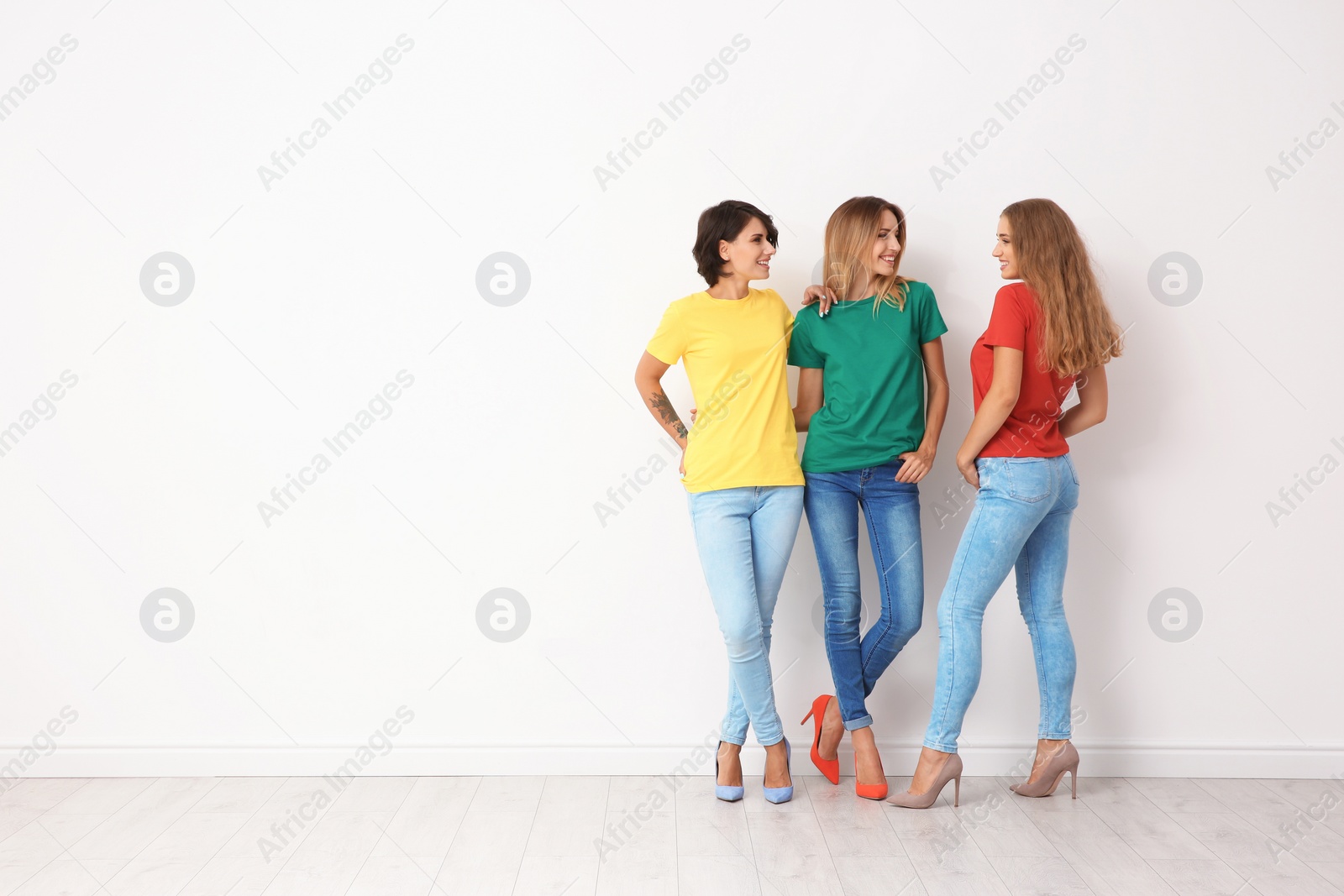 Photo of Group of young women in jeans and colorful t-shirts near light wall