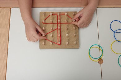 Photo of Motor skills development. Girl playing with geoboard and rubber bands at white table, closeup