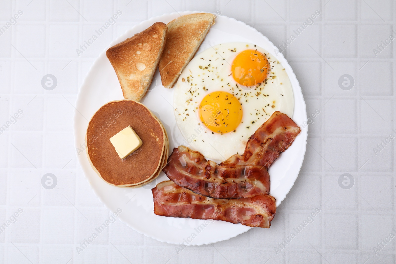Photo of Tasty pancakes with fried eggs and bacon on white tiled table, top view