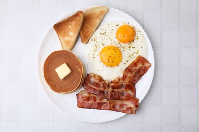 Tasty pancakes with fried eggs and bacon on white tiled table, top view