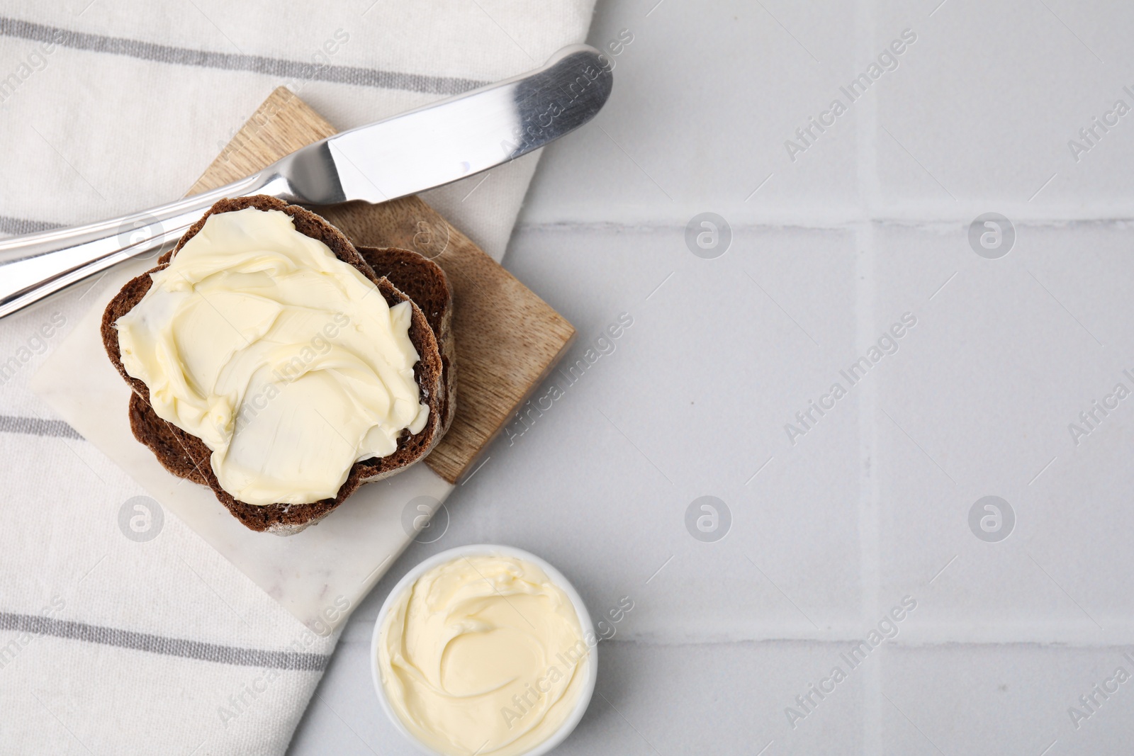 Photo of Slices of tasty bread with butter and knife on white tiled table, flat lay. Space for text