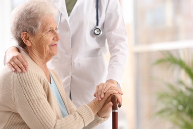 Photo of Nurse assisting elderly woman with cane indoors. Space for text