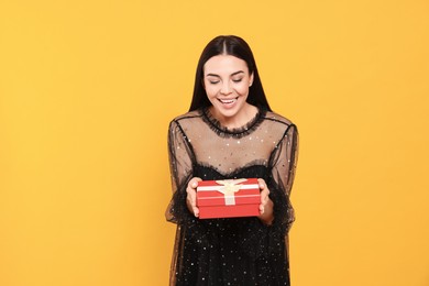 Photo of Happy young woman in festive dress holding gift box on orange background