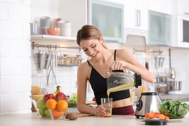 Young woman pouring tasty healthy smoothie into glass at table in kitchen