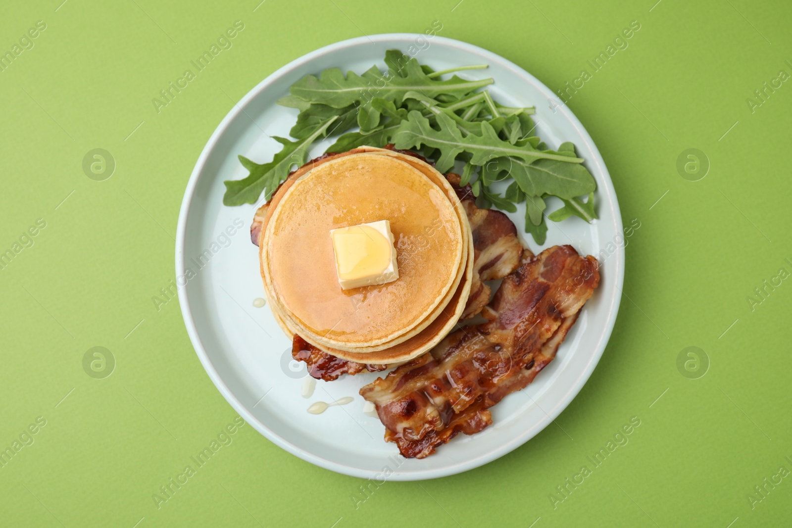 Photo of Tasty pancakes with butter, fried bacon and fresh arugula on light green background, top view