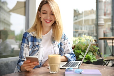 Photo of Young woman using mobile phone while working with laptop at desk in cafe