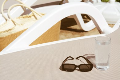 Photo of Stylish sunglasses and glass of water on grey sunbed outdoors, space for text