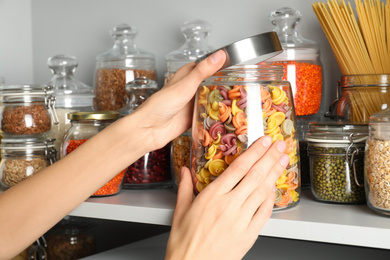 Photo of Woman taking jar of colorful pasta from white shelf, closeup