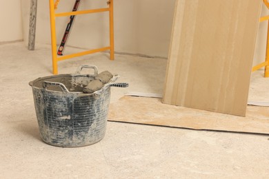 Photo of Bucket of adhesive mix with spatula near tile on floor indoors