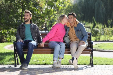 Woman kissing another man while holding hands with her boyfriend on bench in park. Love triangle