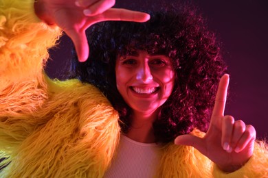 Photo of Beautiful young woman in yellow fur coat making frame gesture with hands on color background in neon lights