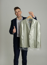 Man holding hanger with jacket in plastic bag on light grey background. Dry-cleaning service