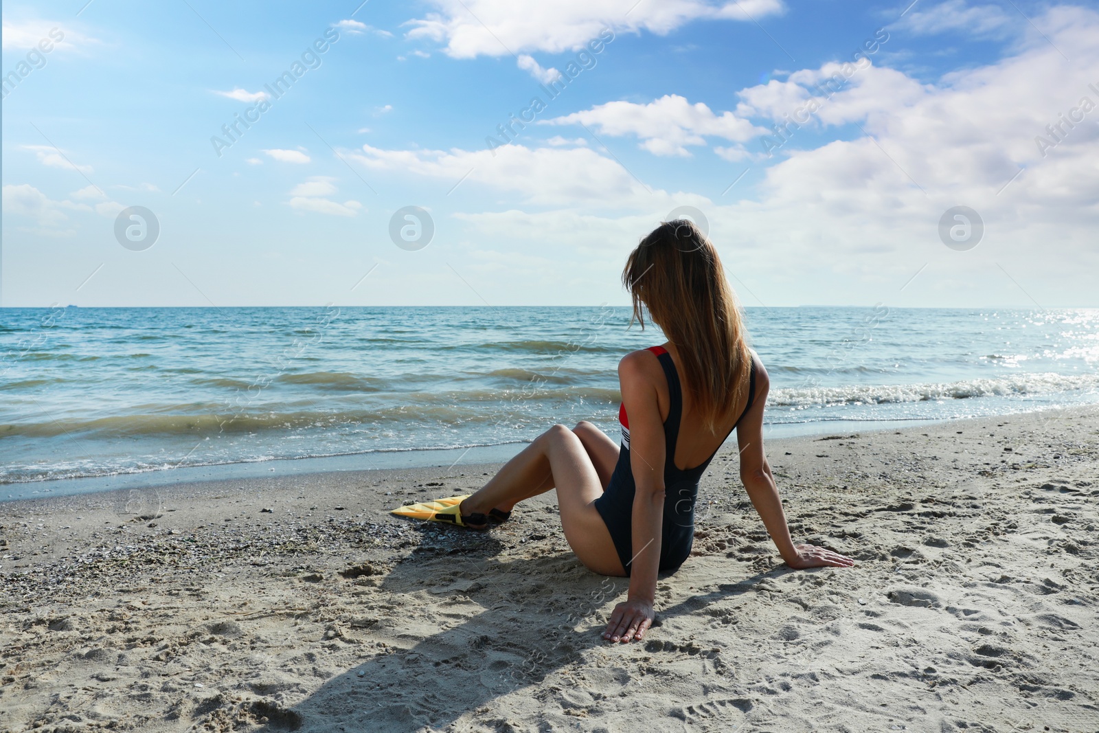 Photo of Woman in flippers sitting near sea on beach, back view