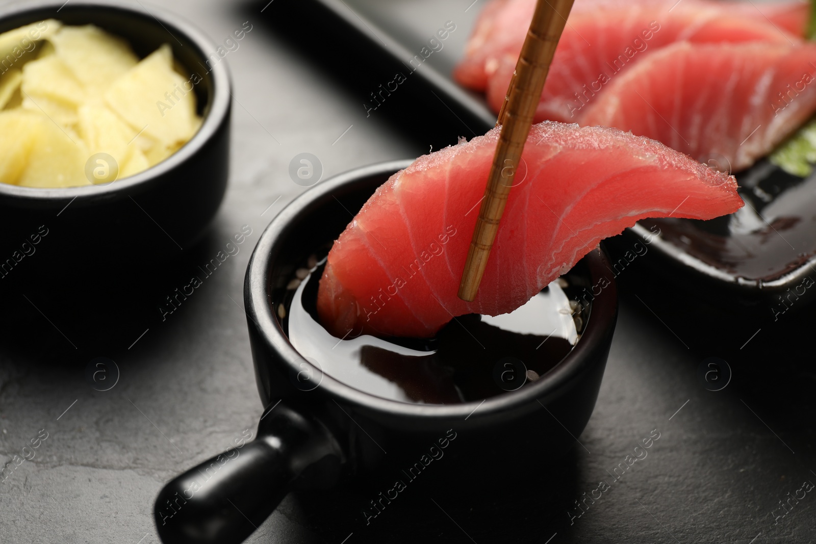 Photo of Dipping tasty sashimi (piece of fresh raw tuna) into soy sauce at black table, closeup