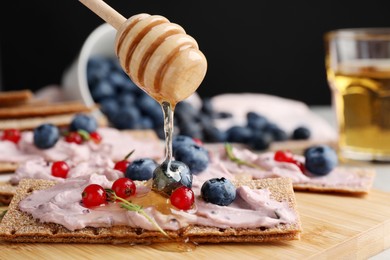 Photo of Pouring honey onto tasty cracker sandwich with cream cheese, blueberries, red currants and thyme on wooden board, closeup