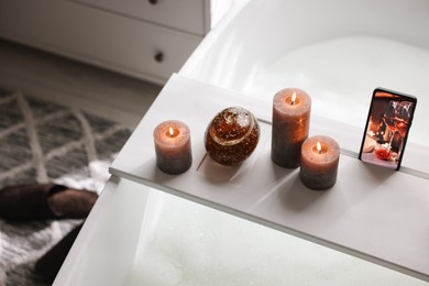 Photo of White wooden tray with smartphone and burning candles on bathtub in bathroom, above view