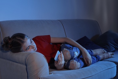Photo of Teenage girl with smartphone lying on sofa in dark room. Danger of internet