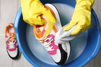 Photo of Woman with gloves and brush cleaning stylish sneakers in wash basin, top view