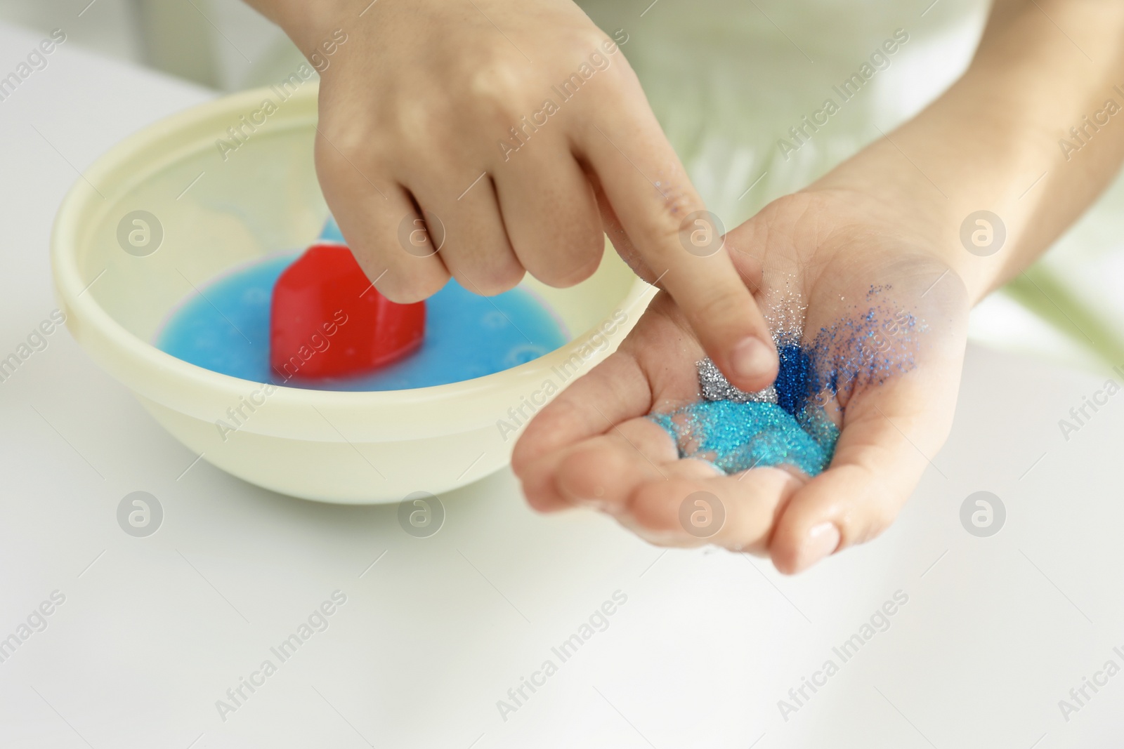 Photo of Little girl adding colored sparkles to homemade slime toy at table, closeup