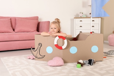 Photo of Cute little girl playing with cardboard ship at home