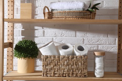 Photo of Toilet paper rolls in wicker basket, floral decor and cotton pads on wooden shelf against white brick wall