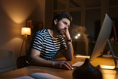 Photo of Home workplace. Tired man working with computer at wooden desk in room at night